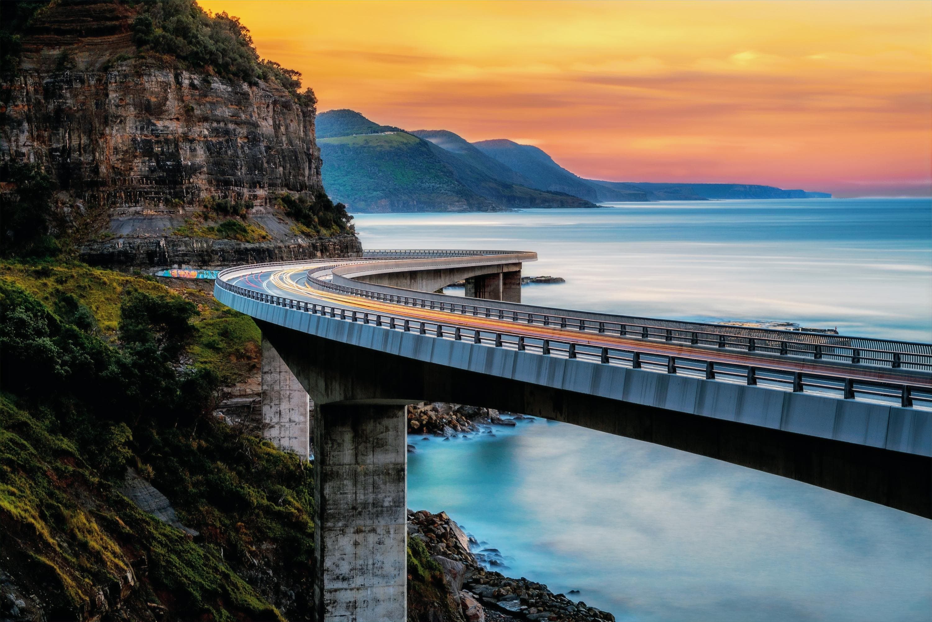 The Sea Cliff Bridge curving along the Australian coastline at sunset, a road leading onward